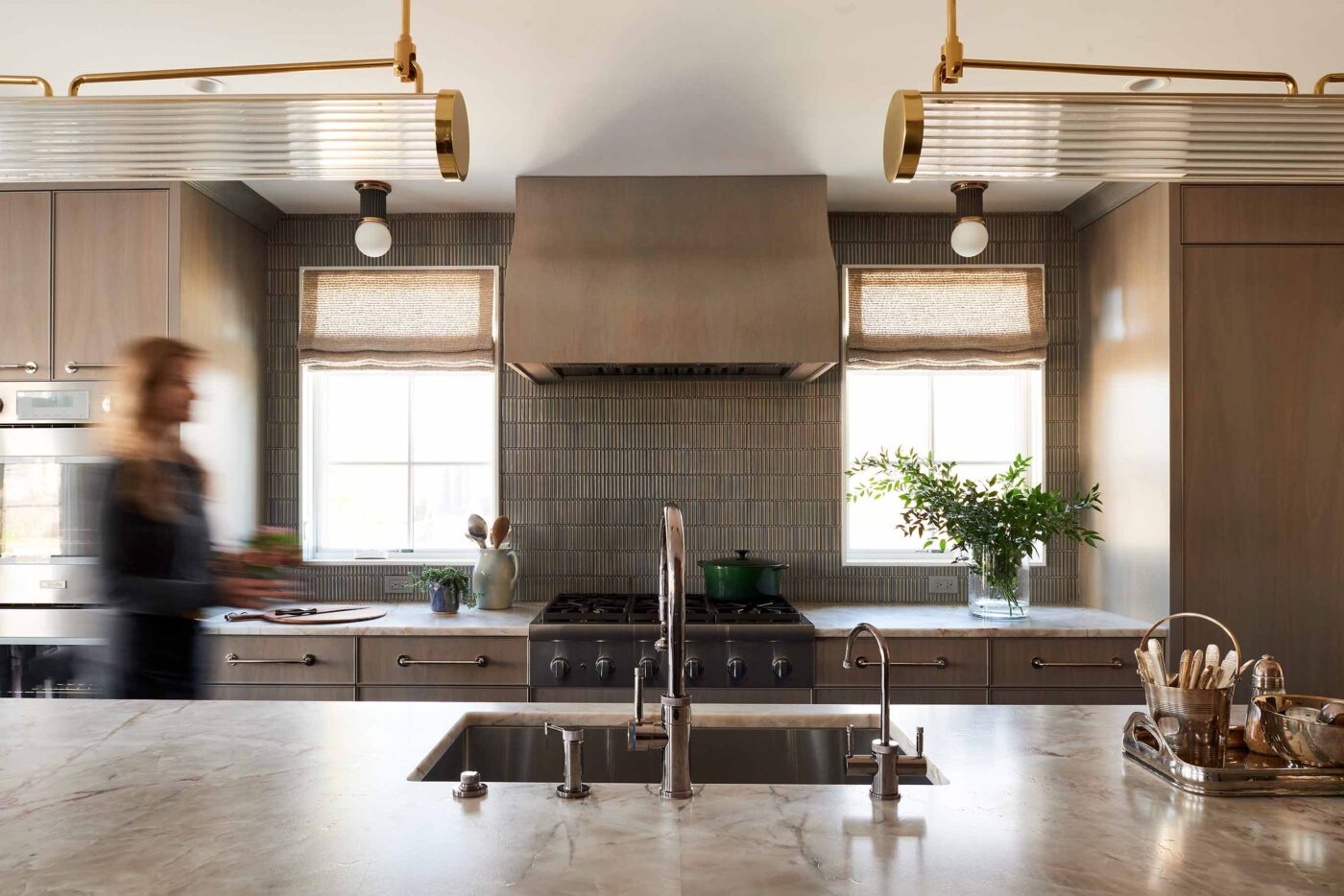 This image shows a modern kitchen featuring light grey cabinets, quartz countertops, custom, curved wood vent hood, light blue ribbed tile, polished nickel hardware, polished nickel plumbing fixtures.View of the cooktop and island sink.