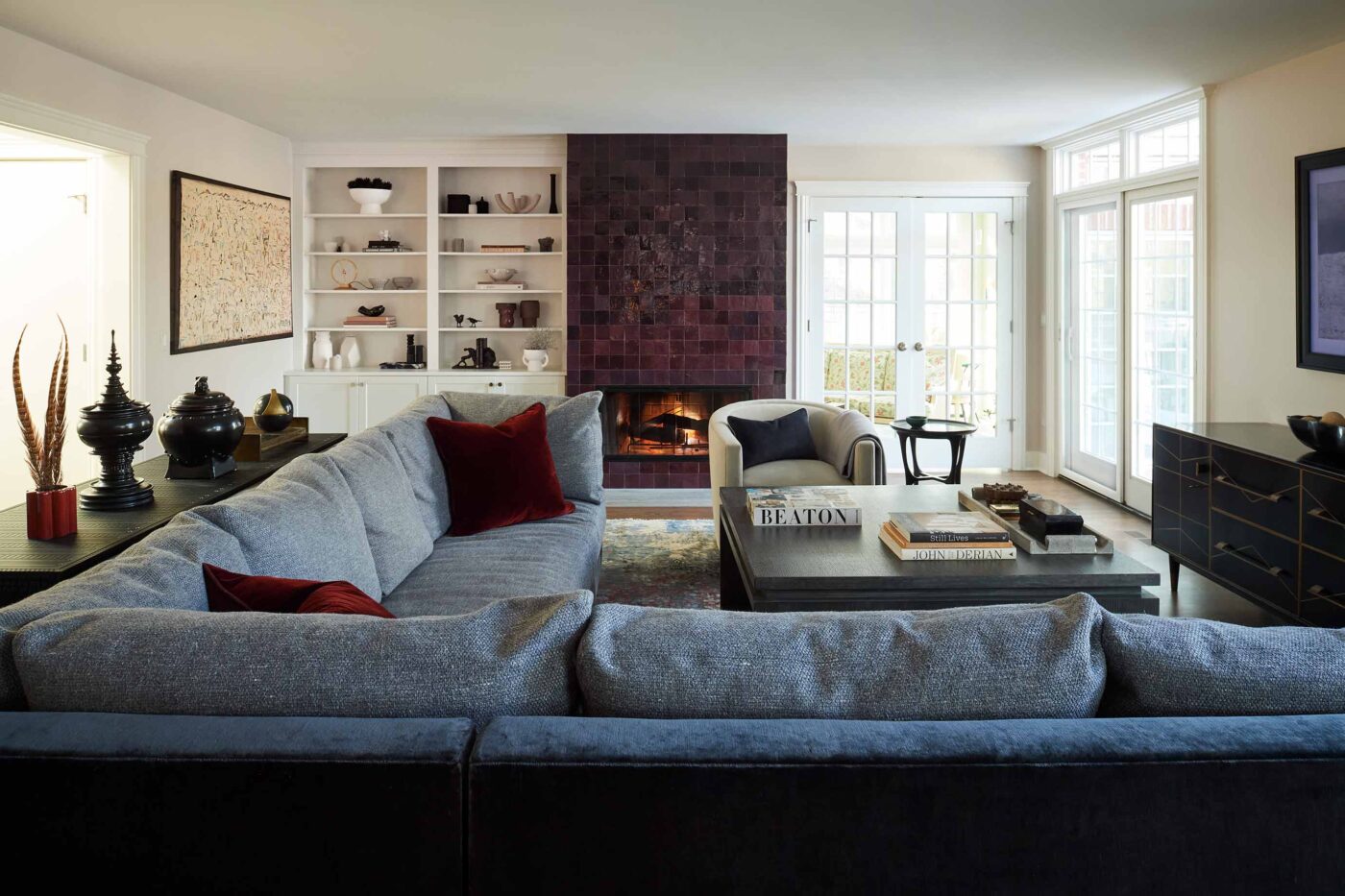 this image is shows a family room with a blue and grey sectional sofa; deep burgundy, wine colored Moroccan Zelige tiles, white bookshelves, dark wood tables